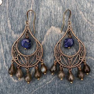 bronze chandelier earrins with five labradorite drop beads. the center has a lapis lazuli carved skull hanging with jump rings. brass ear wires. background is a dark blue stained wood.